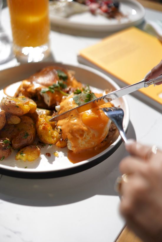A person is cutting into a poached egg on a plate with roasted potatoes, with a beverage and another dish in the background.