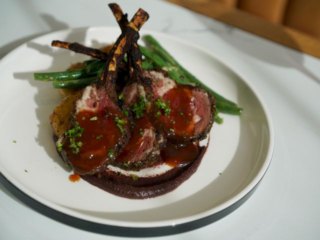 The image shows a plated dish of lamb chops with a sauce, green beans, and garnished with herbs, on a white plate.
