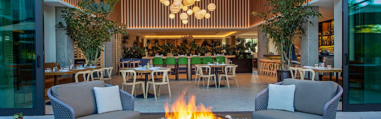 An outdoor seating area with a fire pit and modern chairs, leading into a stylish restaurant with wooden accents and hanging lights.