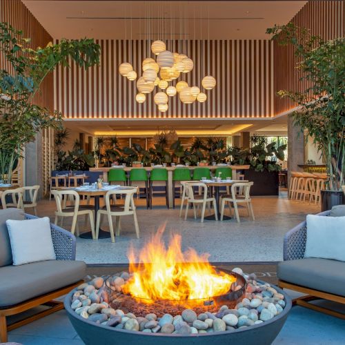 An outdoor seating area with a fire pit and modern chairs, leading into a stylish restaurant with wooden accents and hanging lights.