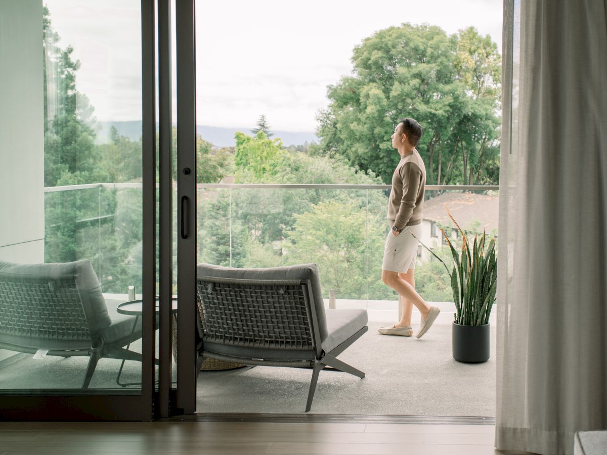 A person walks on a balcony with outdoor furniture and a potted plant, overlooking greenery and trees.