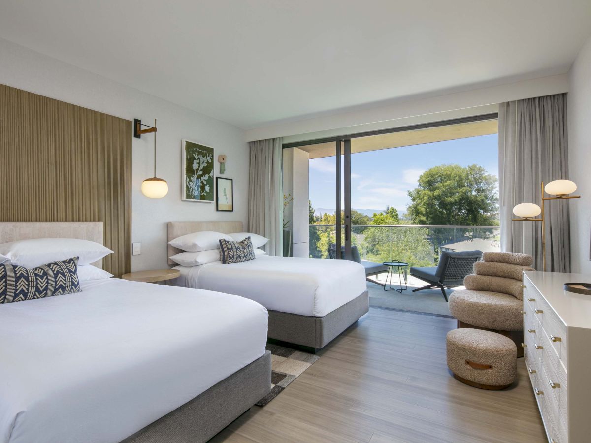 A modern hotel room with two beds, stylish decor, and a balcony with a view of trees and outdoor furniture.