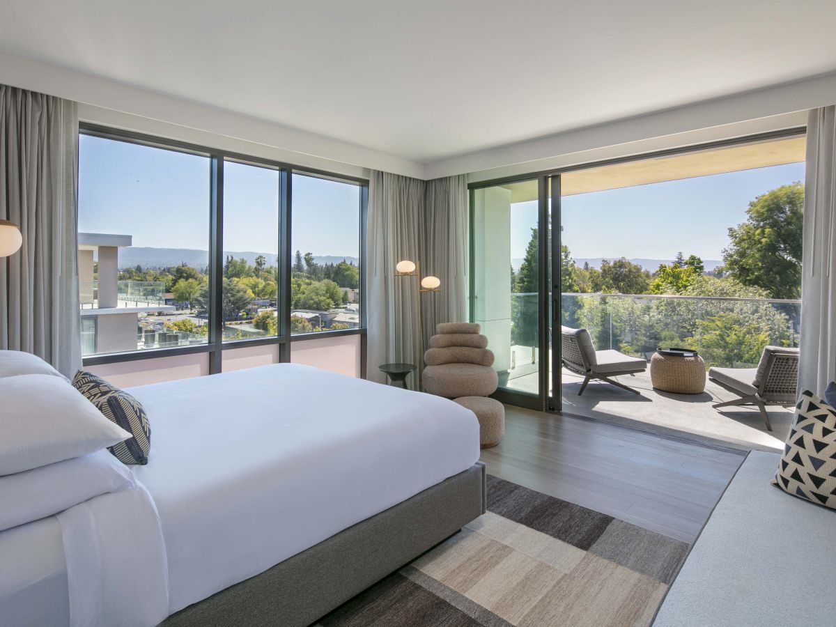 A modern hotel room with a bed, unique chair, floor lamp, and balcony view of trees and buildings outside.