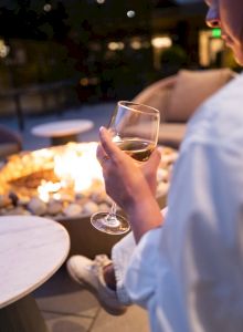 A person holding a glass of wine sits by an outdoor fire pit, with a cozy seating setup in the background.