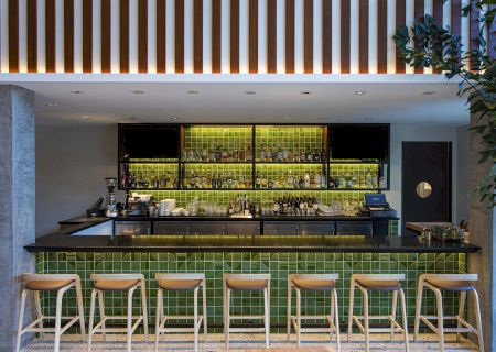 A modern bar with green tiled backsplash, stocked shelves, wooden stools, and a sleek countertop, lit with overhead lighting.