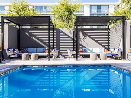 A swimming pool with cabanas, lounge chairs, and tables surrounded by trees and a modern building in the background.