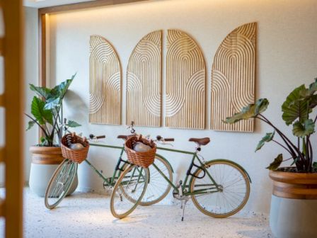 Two bicycles with baskets stand against a wall with geometric art, surrounded by large potted plants.