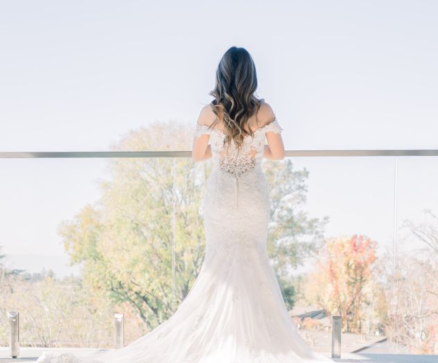 A person in a white wedding dress stands on a balcony, overlooking a scenic view with trees.