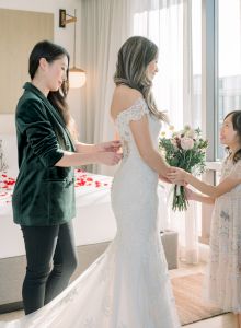 A bride in a white dress is helped by two women in a sunlit room, one adjusting the dress, one holding flowers.