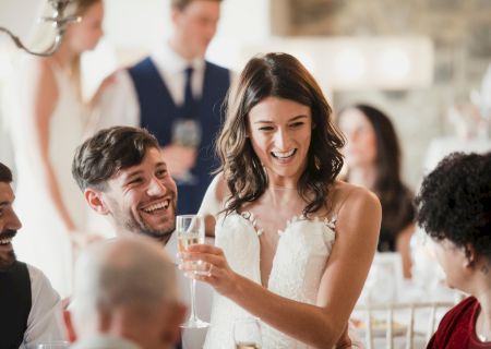 A group of people celebrates at a wedding, with a bride and a man smiling and holding champagne glasses, enjoying the joyful moment.