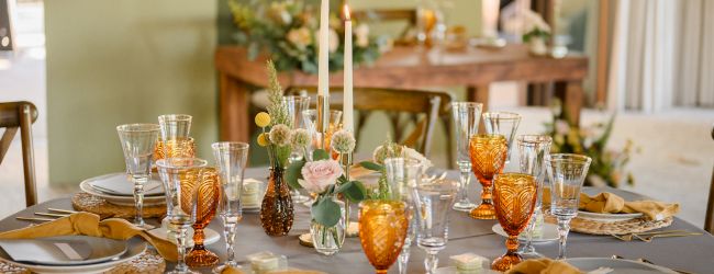 A beautifully set dining table with elegant glassware, orange goblets, candles, and floral centerpieces, creating a warm ambiance.