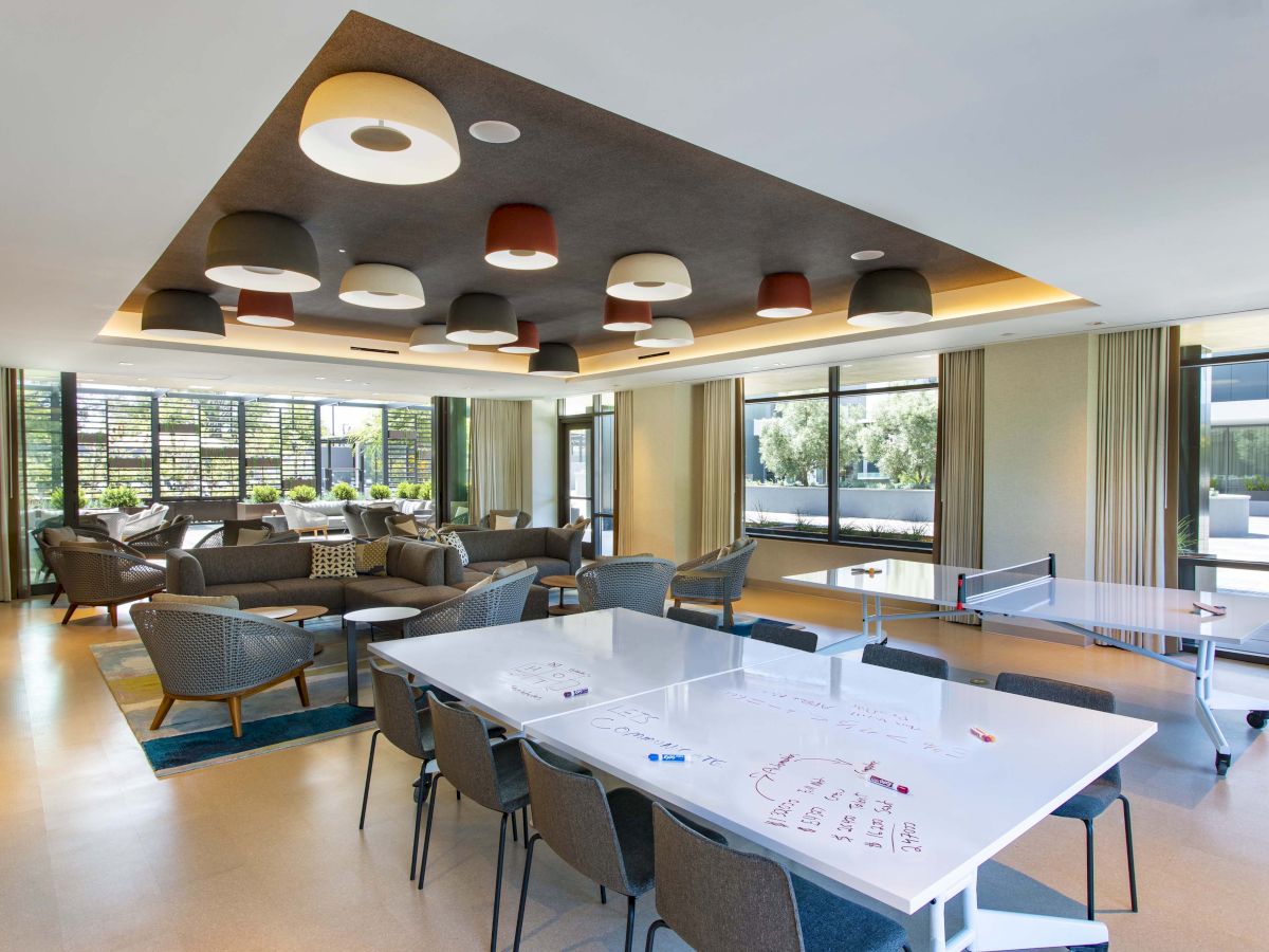 The image shows a modern lounge area with comfy seating, a table tennis setup, and large windows, featuring unique ceiling lights.