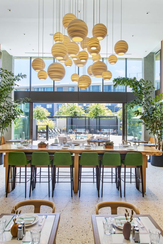 A modern restaurant with wooden decor, green chairs, and hanging lights, featuring large windows and potted plants.